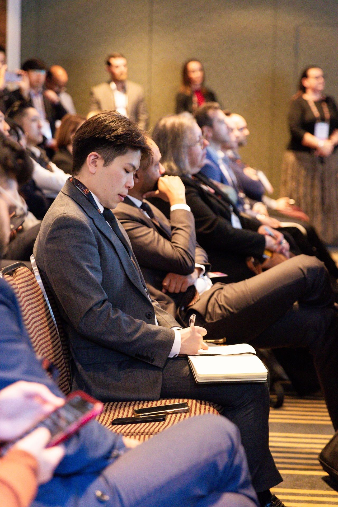 Audience seated at a conference, person in the foreground taking notes, others listening attentively.