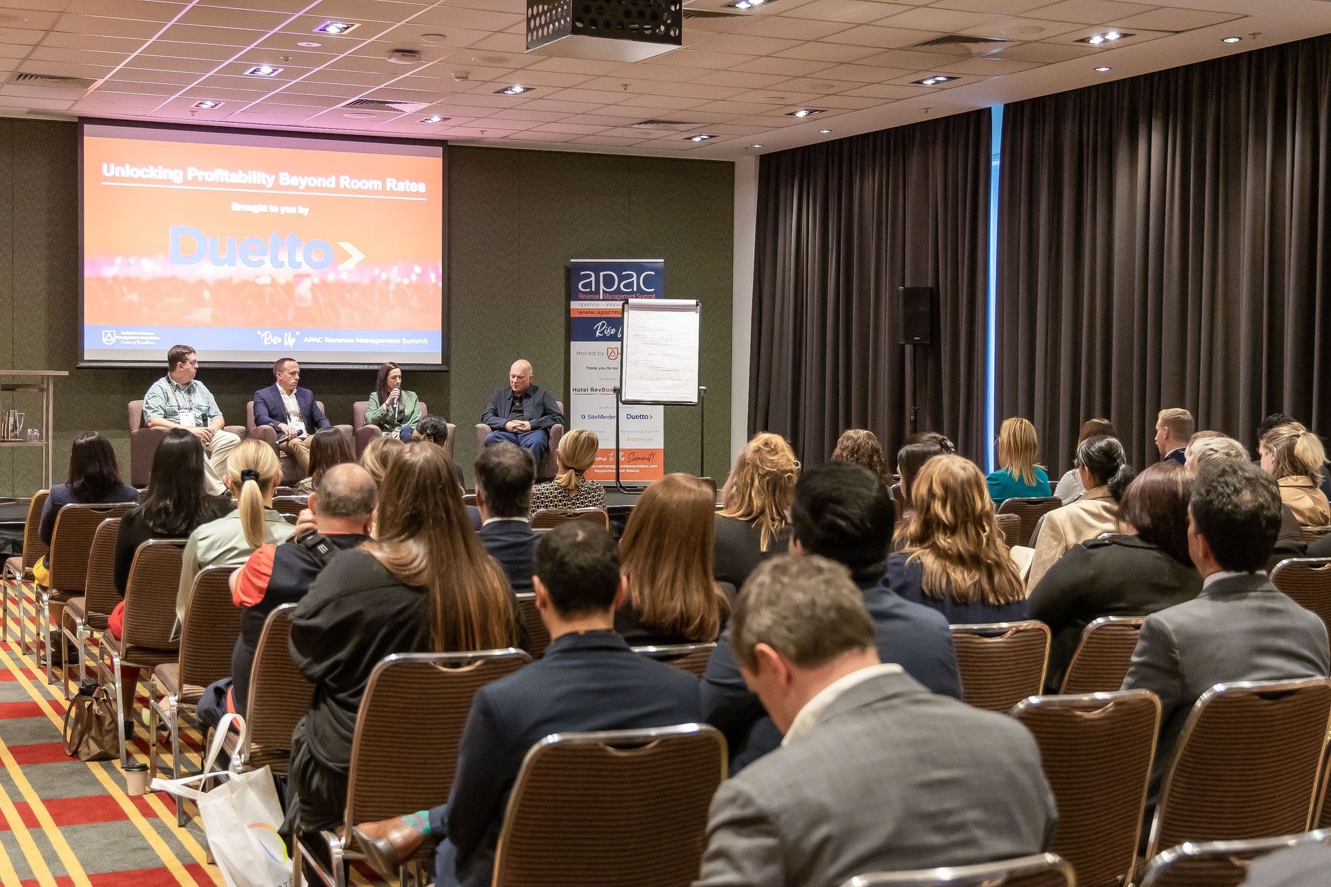 Panel discussion at a conference with four speakers seated on stage and audience watching.